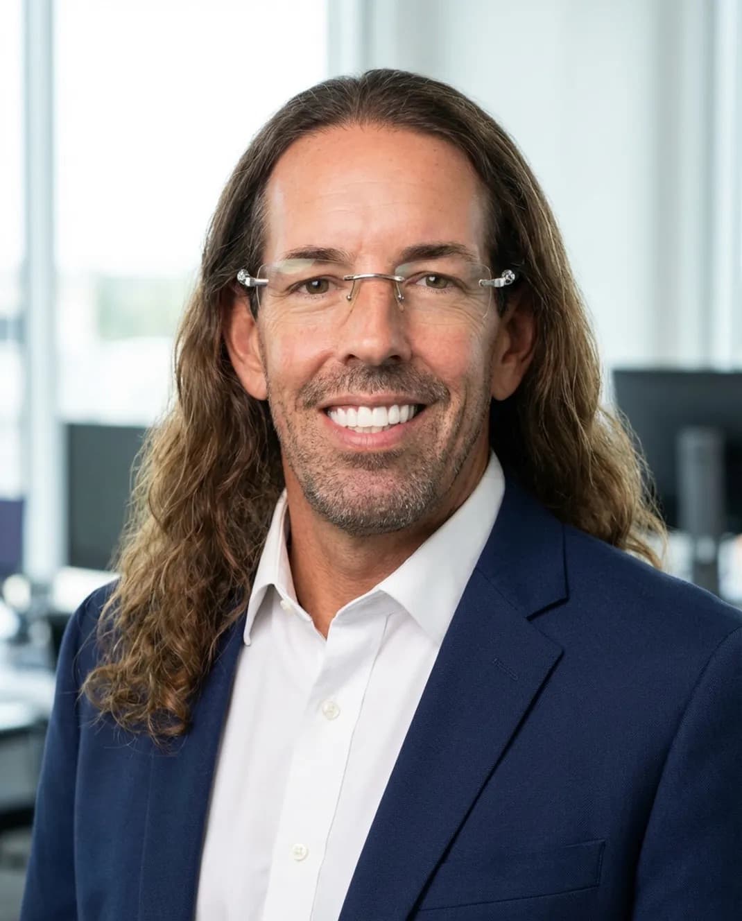 Smiling man with long wavy hair and rimless glasses wearing blue blazer and white shirt.