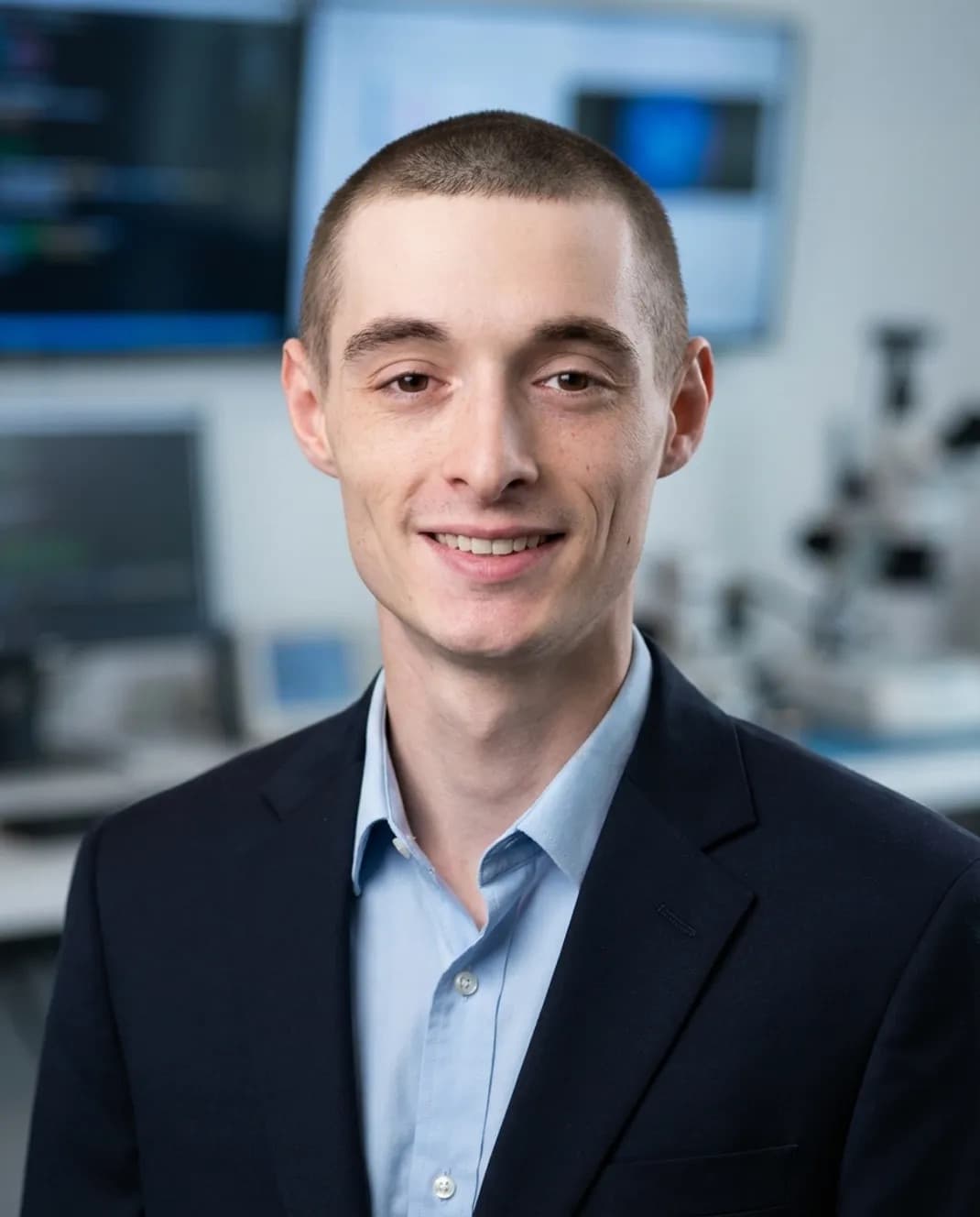 Smiling young man with short hair wearing a navy blazer and light blue shirt.